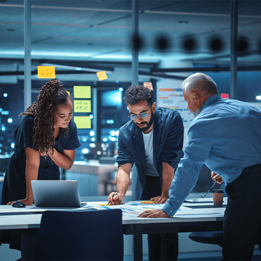 A diverse team collaborates at a brightly lit conference table, discussing ideas on paper and a laptop against a city night backdrop.