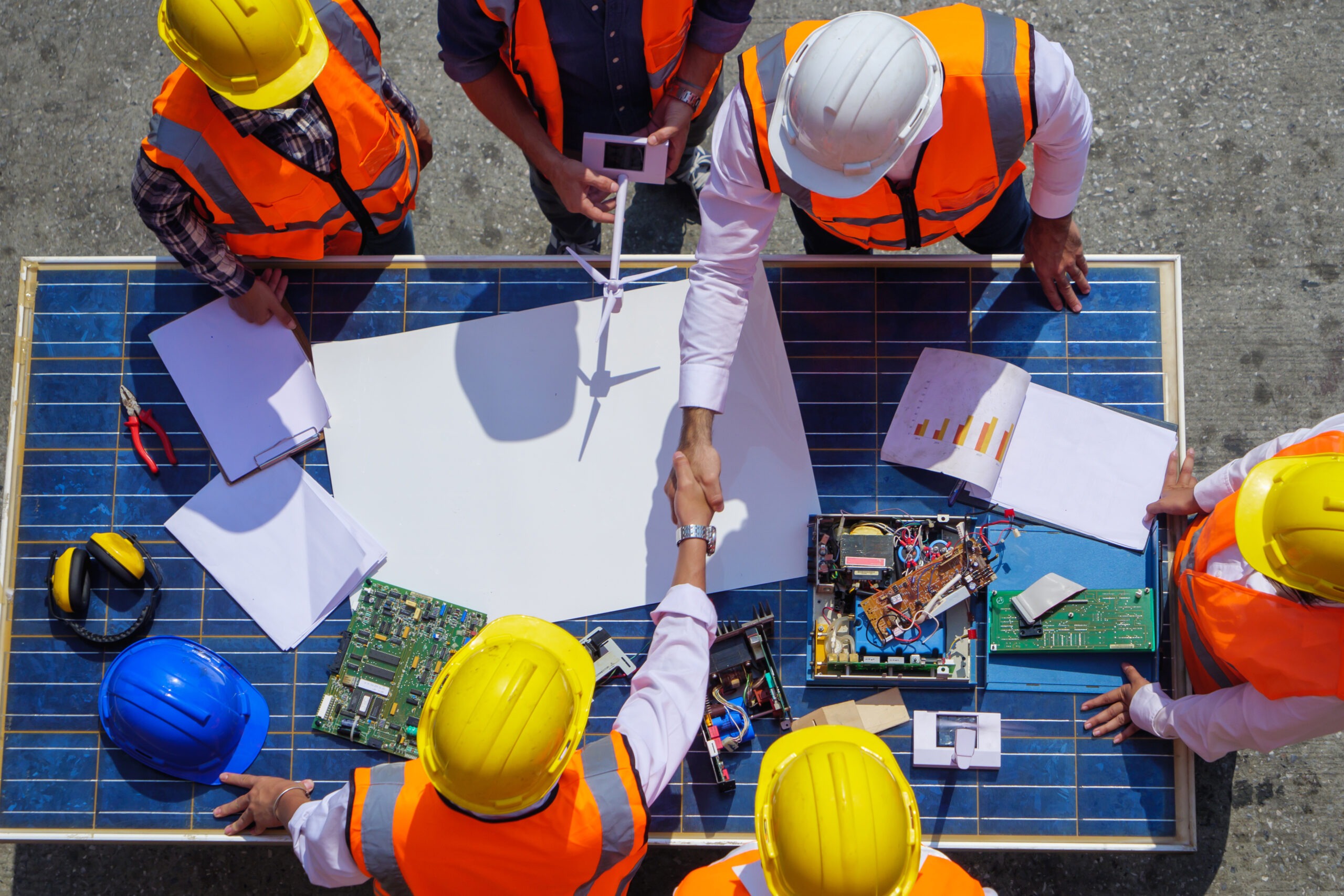 Engineers and technicians in safety gear discuss plans and collaborate over a table with solar panels and electronic components.