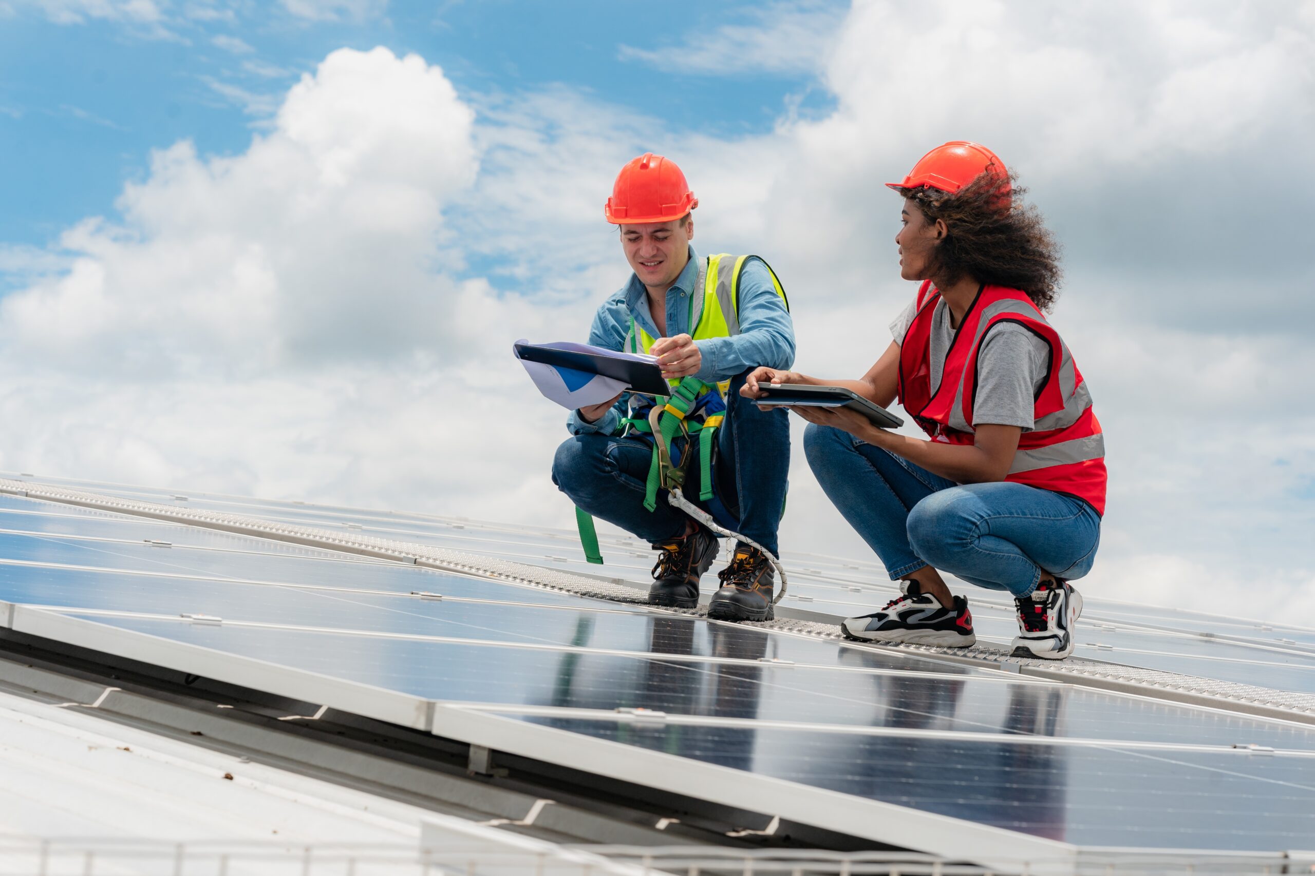 Two technicians, wearing safety helmets and harnesses, inspect solar panels on a rooftop while discussing plans and taking notes under a partly cloudy sky.